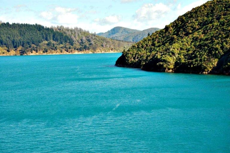 A serene view of a turquoise bay surrounded by lush green hills and mountains, with a bright sky and scattered clouds above