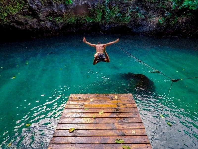 A person diving into a clear blue natural pool from a wooden platform, surrounded by lush greenery.