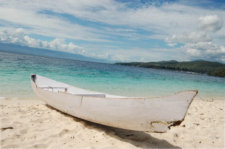 Visit Tanjung Karang Beach for an idyllic paradise, white wooden rowing boat sitting on sandy beach with turquoise waters of the ocean behind under a cloudy blue sky