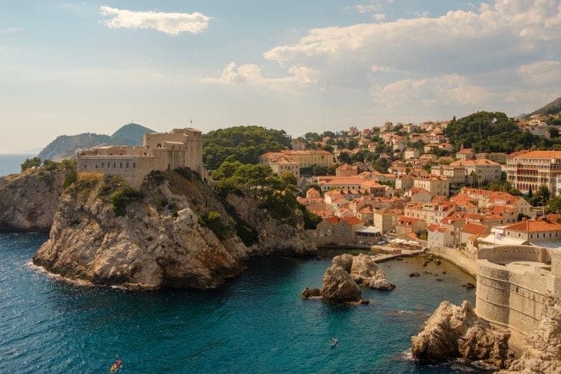 Enjoy this Dubrovnik guide for non Game of Thrones fans, view of harbour of Dubrovnik with tall rocky hills on one side and a densely packed built up area on the other with many buildings with terracotta rooftops lining the rolling hills interspersed with green trees all under a bright sky with white clouds