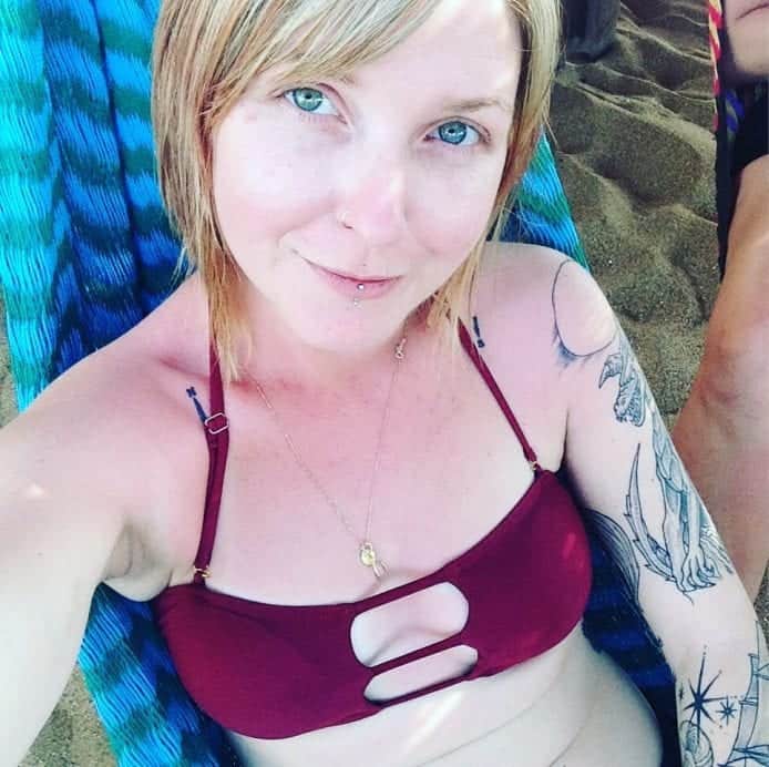Selfie of blonde woman with blue eyes and tattoos on her left arm wearing a bathing suit and sitting in a hammock on the beach