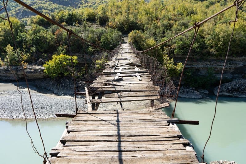 Discover unique things to do in Albania, view looking out across wooden suspension footbridge in moderate state of disrepair hanging over river with green trees on the opposite side