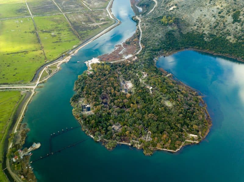 Visiting Butrint National Park, aerial shot of Butrint National Park with ruins and ancient buildings nestled amongst trees and green areas all surrounded by clear blue water