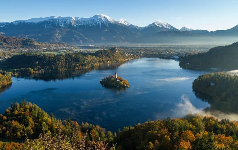 Discover the best things to do in Slovenia for free, majestic view of small island on which stands a small building with pointed tower in the centre of a clear lake surrounded by forest and fields with large range of snow-capped mountains in the background