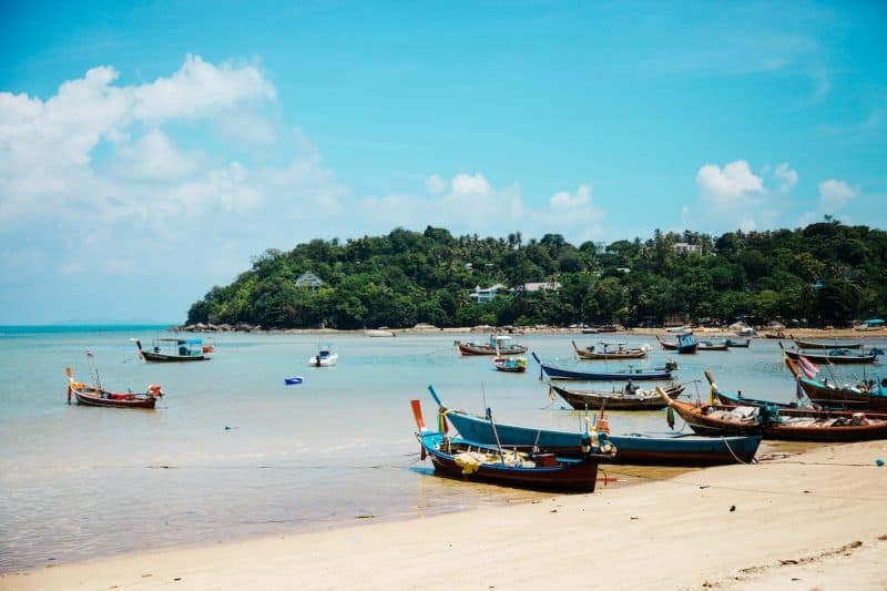 Find the best ways for how to get to Koh Samui, beach with small boats moored on the shore and in the shallows with a large peninsula covered in green trees behind under a bright blue sky with some white clouds