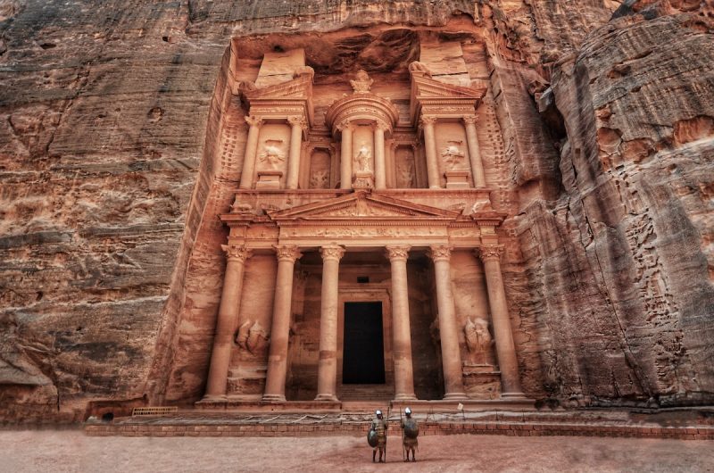 Enjoy visiting Petra this year, view of large stone entranceway covered in ornate statues and pillars carved into the side of an enormous stone cliff with two people in traditional dress standing in front