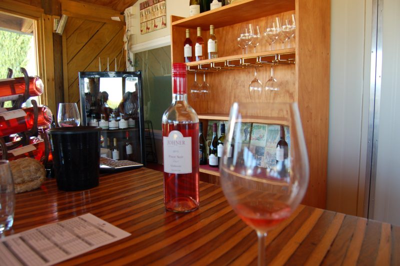 A cozy wine tasting room with a focus on a bottle of Johner 2015 Pinot Noir Rosé placed on a wooden countertop. The glass in the foreground contains a small amount of the rosé wine, catching the light beautifully. Behind the counter, shelves display more wine bottles and hanging wine glasses, while a wine rack on the left holds several bottles of rosé. The warm wood tones of the room, combined with the sunlight filtering through the windows, create an inviting and intimate atmosphere for wine tasting.