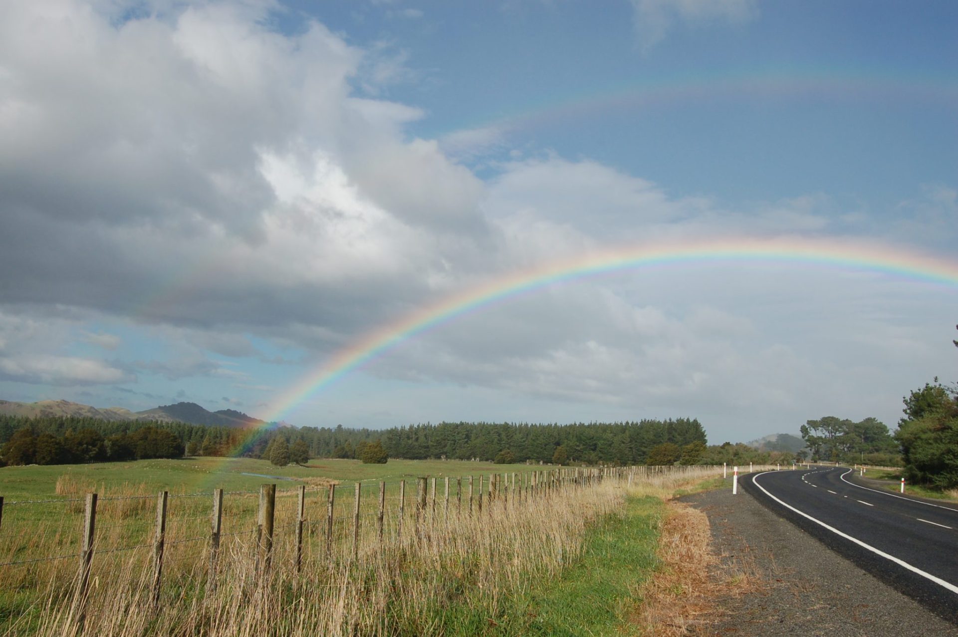 A peaceful countryside scene with a two-lane road stretching alongside a grassy field. A double rainbow arcs across a partly cloudy sky, adding vibrant colors to the landscape. A wooden fence lines the left side of the road, separating it from the open fields, while trees and distant hills are visible in the background. The atmosphere feels serene and calm, with the road leading toward the horizon.