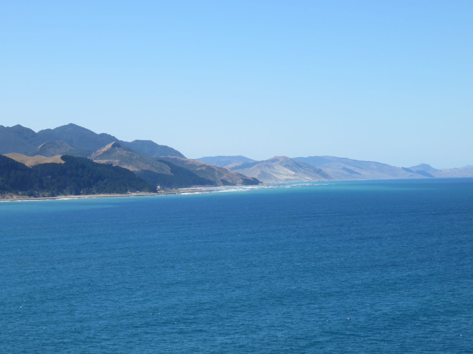 A coastal landscape with a deep blue ocean in the foreground and a range of mountains stretching along the shoreline under a clear, blue sky. The mountains taper off into the distance, with lighter-colored sandy slopes visible near the shoreline.
