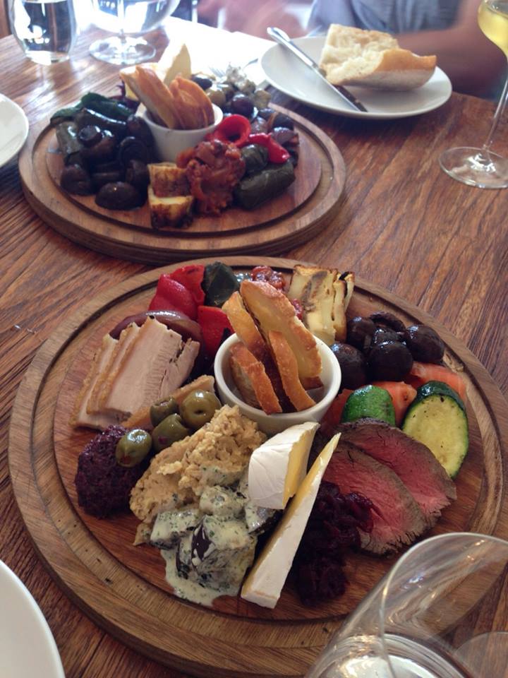 A round wooden platter filled with assorted appetizers, including sliced meats, cheeses, grilled vegetables, olives, hummus, and breadsticks, arranged artfully on a rustic table.