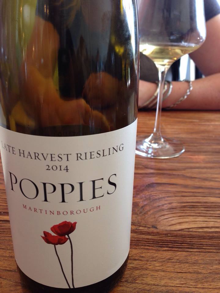 A bottle of Poppies Late Harvest Riesling 2014 from Martinborough, labeled with a red poppy flower illustration, stands on a wooden table next to a glass partially filled with white wine.