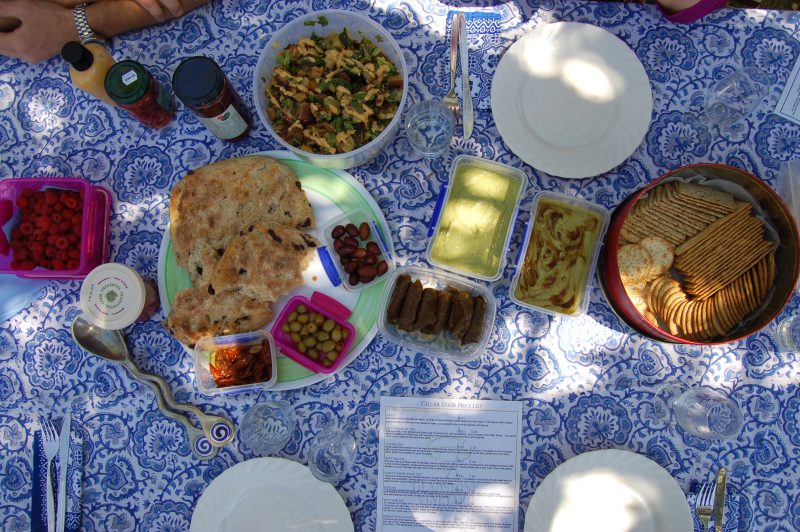 An overhead shot of a picnic table filled with food, including bread, olives, salad, and dips, set on a blue-patterned tablecloth in an outdoor setting.