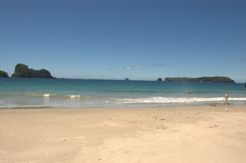 This image shows a serene beach scene with golden sand in the foreground and gentle waves rolling onto the shore. The turquoise ocean stretches out to distant, lush green islands under a clear blue sky, with a few people enjoying the water and the shoreline.