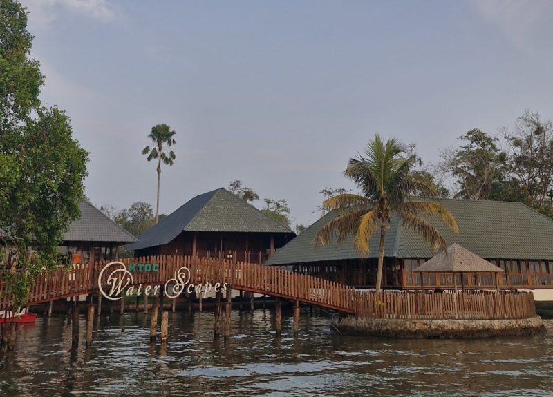 The entrance to "KTDC Water Scapes," a stunning resort with wooden walkways extending over a serene lake, surrounded by lush tropical trees.